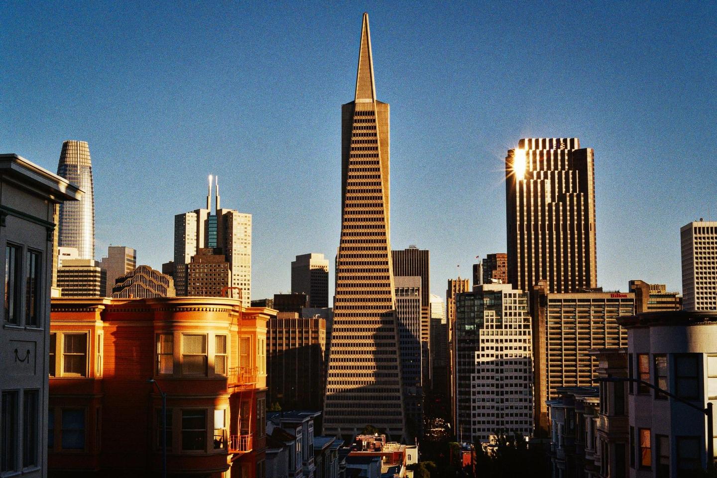 A cityscape with a prominent triangular skyscraper and other buildings is illuminated by golden hour sunlight.