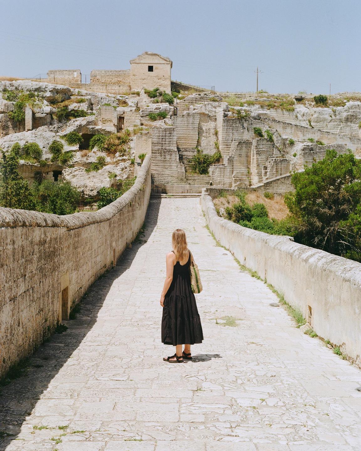A woman stands on a stone bridge looking towards a hillside with ancient stone structures and vegetation.