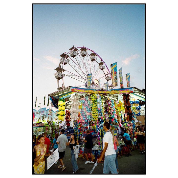 A Ferris wheel rises above a carnival game booth filled with stuffed animals, with people walking in front.