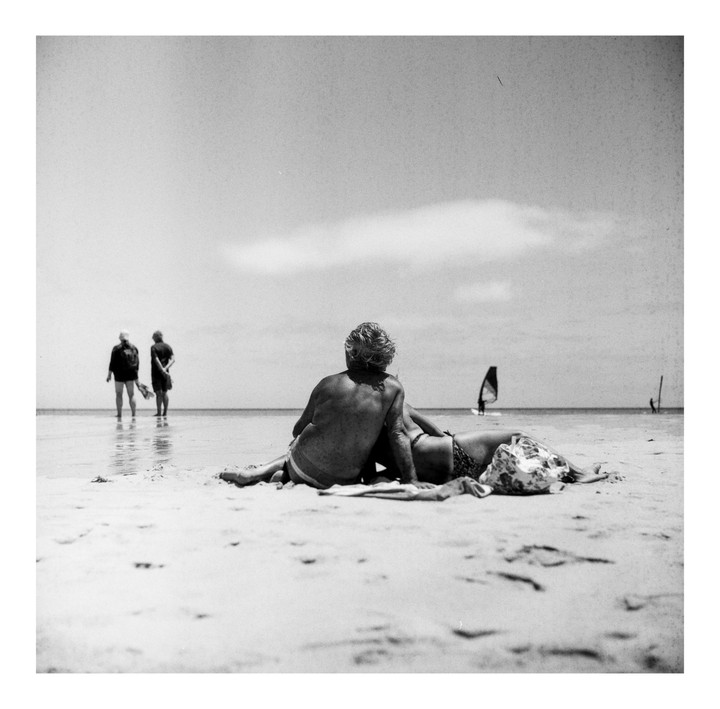 A couple sits on a sandy beach looking out at the water where two people stand and a windsurfer is visible.