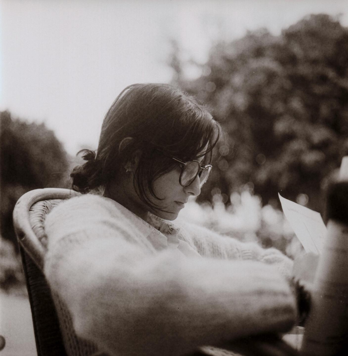 A woman with glasses and a sweater sits in a wicker chair, looking down at a piece of paper.