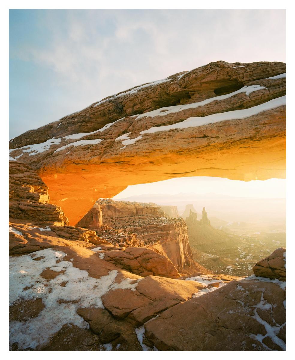 A natural arch frames a canyon landscape with snow-dusted rocks and a bright sunrise.