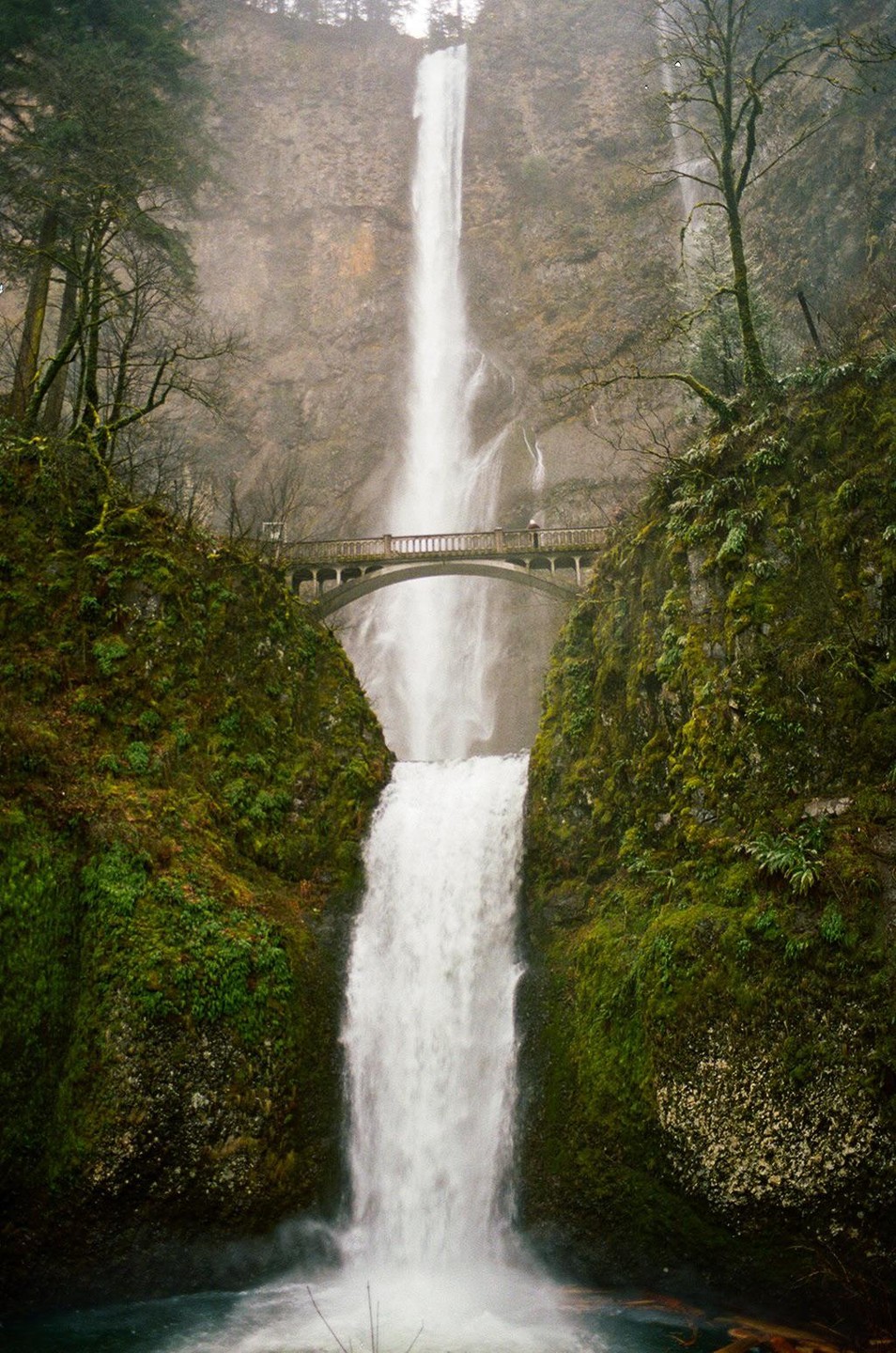 A tall waterfall cascades down a cliff face, with a bridge spanning the middle section of the falls.