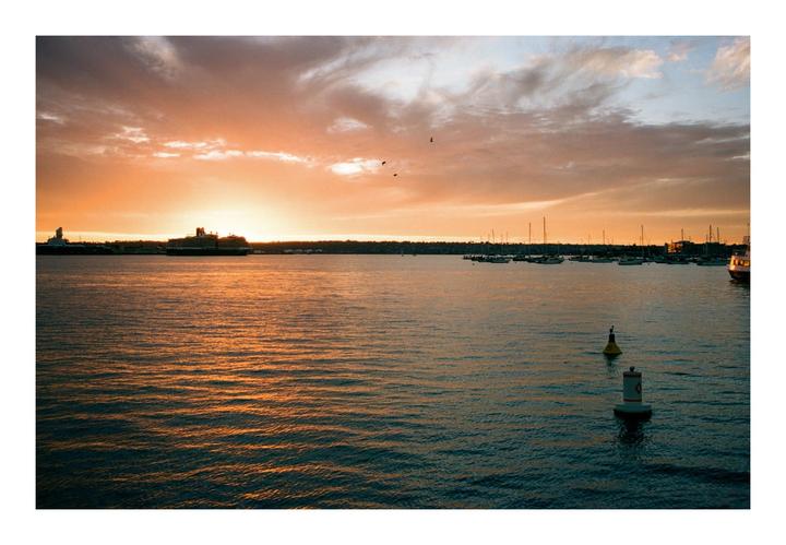 A large body of water with boats and buoys is illuminated by the setting sun, which casts an orange glow across the sky and water.