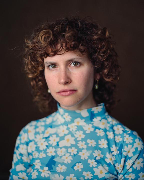 A woman with curly hair wears a blue floral top and looks directly at the viewer.