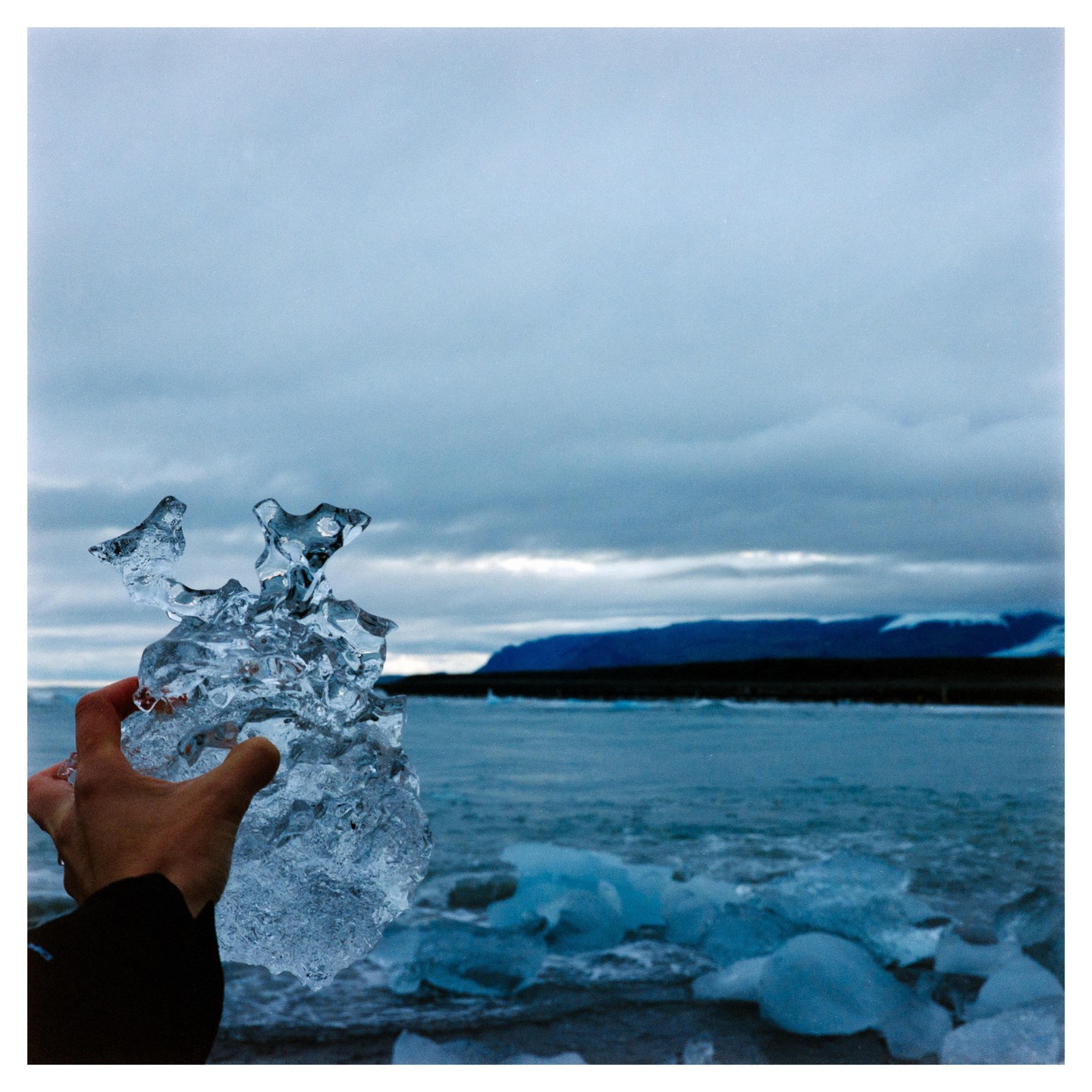 A hand holds a piece of ice in front of a body of water with ice chunks and a distant mountain range under a cloudy sky.