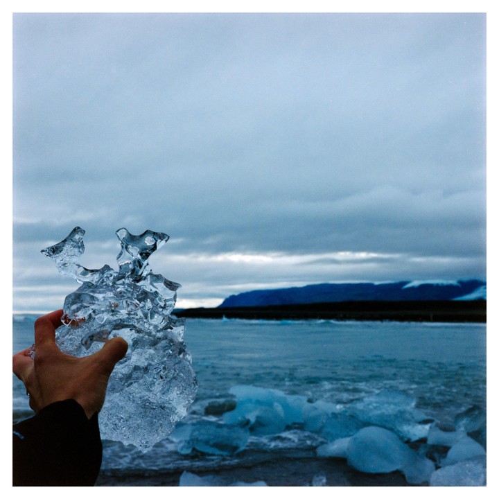 A hand holds a piece of ice in front of a body of water with ice chunks and a distant mountain range under a cloudy sky.