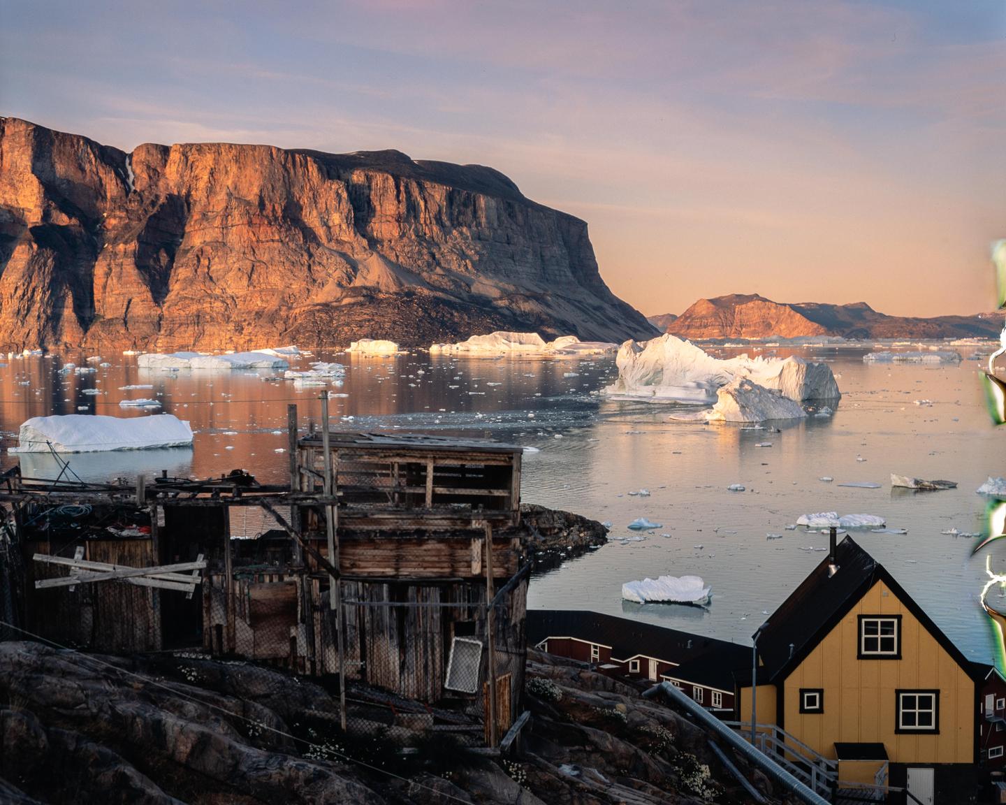 A small settlement with wooden houses sits on a rocky shore, facing a body of water filled with icebergs and backed by a large mountain range under a golden hour sky.