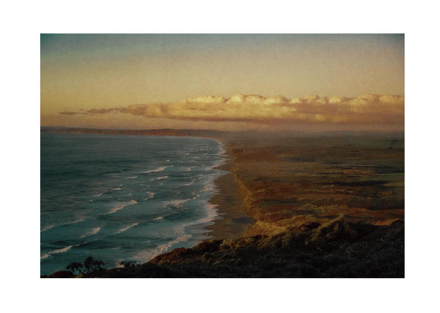 A long stretch of coastline with waves breaking on the shore is visible from a high vantage point under a cloudy sky.