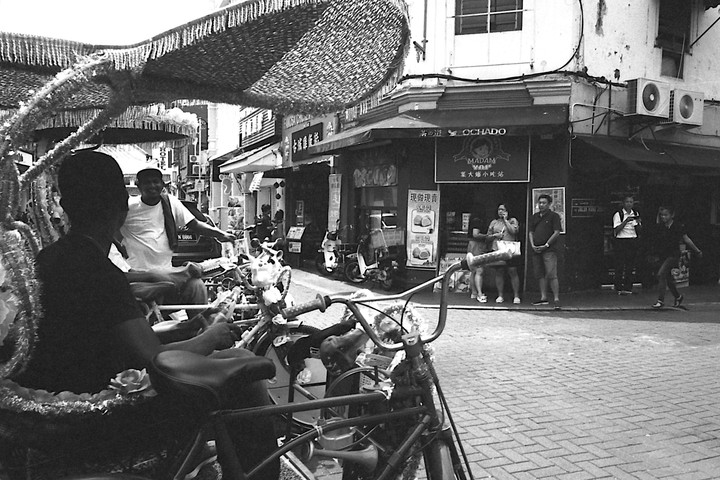 A man sits in a rickshaw on a street with people and shops in the background.