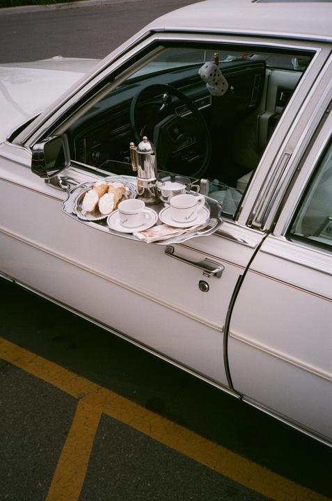 A silver tray with coffee cups and pastries is placed on the open window of a white car.