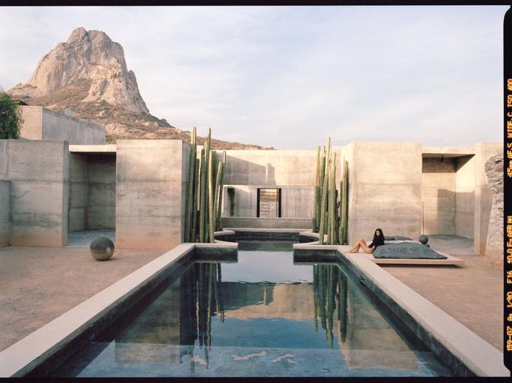 A woman reclines by a pool in a concrete courtyard with cacti, a mountain in the background.