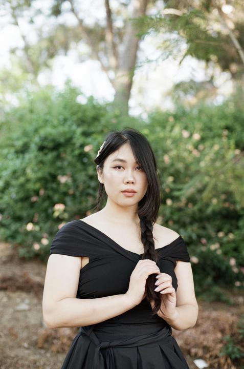 A woman with a braid and a pearl hair clip poses in a black dress outdoors.