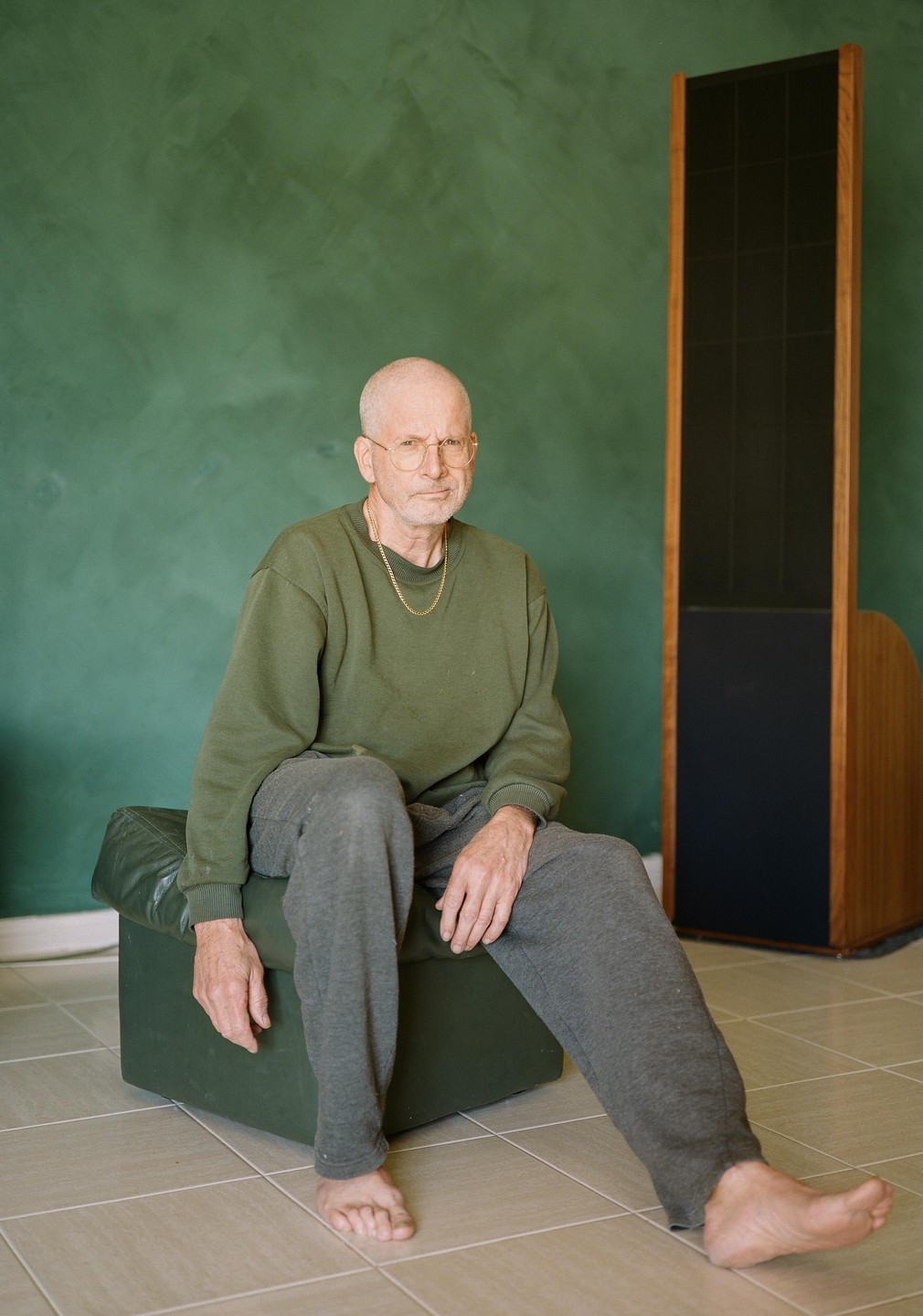 A man wearing glasses, a green sweatshirt, and gray sweatpants sits barefoot on a green ottoman in a room with a green wall and a tall speaker.