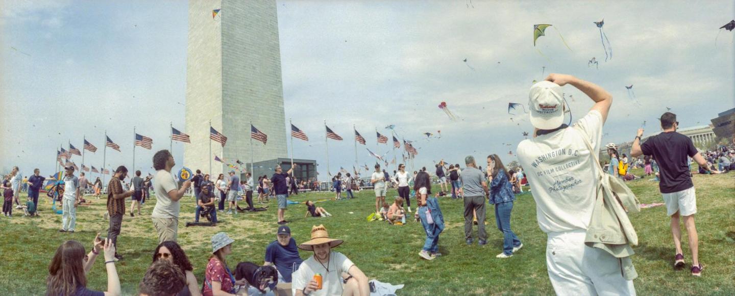 A large crowd of people are gathered on a grassy field with the Washington Monument and many American flags in the background, and kites flying in the sky.