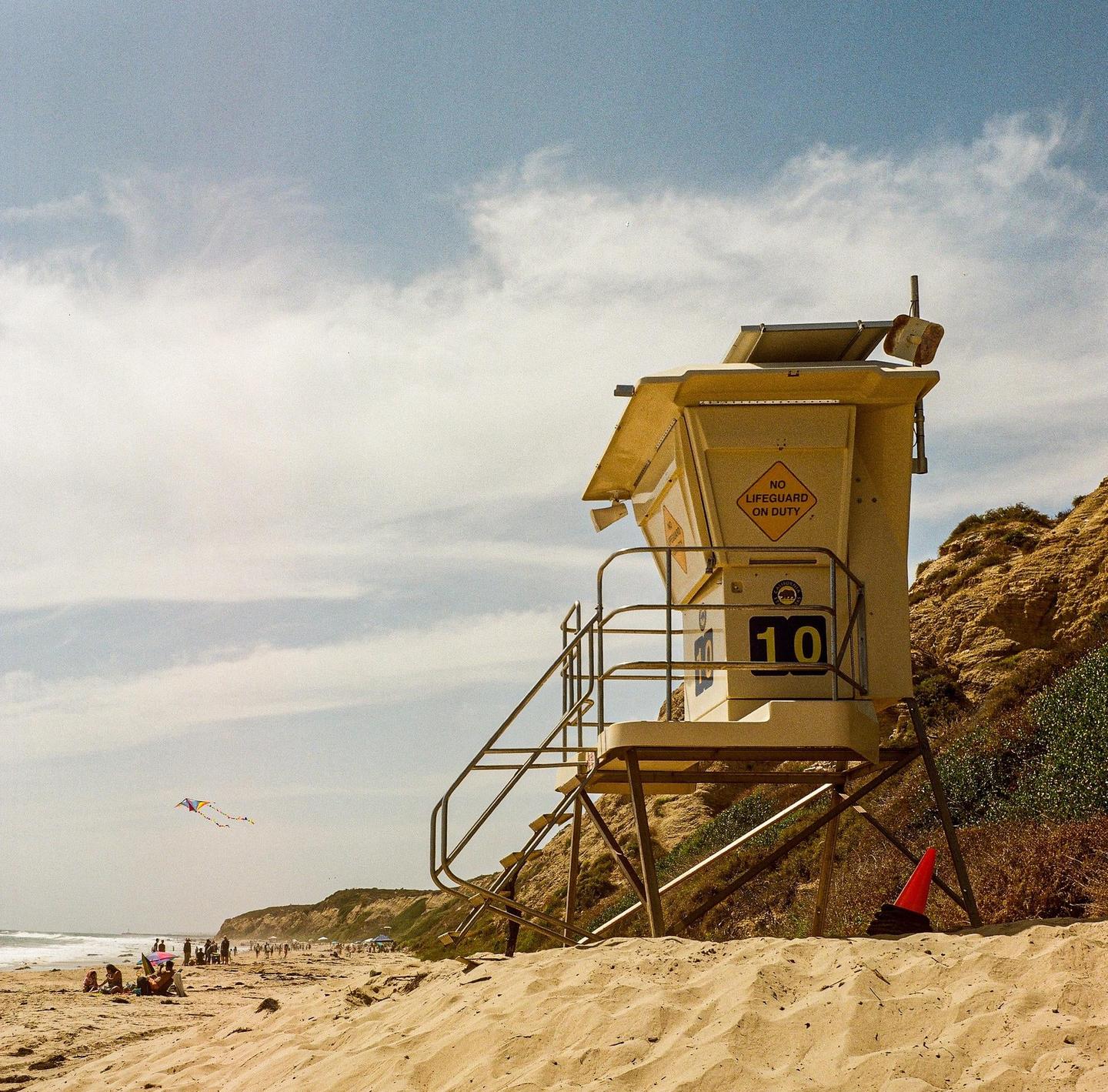A lifeguard tower stands on a sandy beach with people and a kite in the distance.
