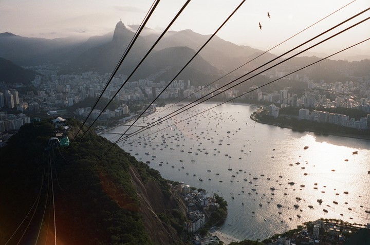 A high-angle view of a bay filled with boats, a city, and mountains in the distance, with cable car wires crossing the frame.