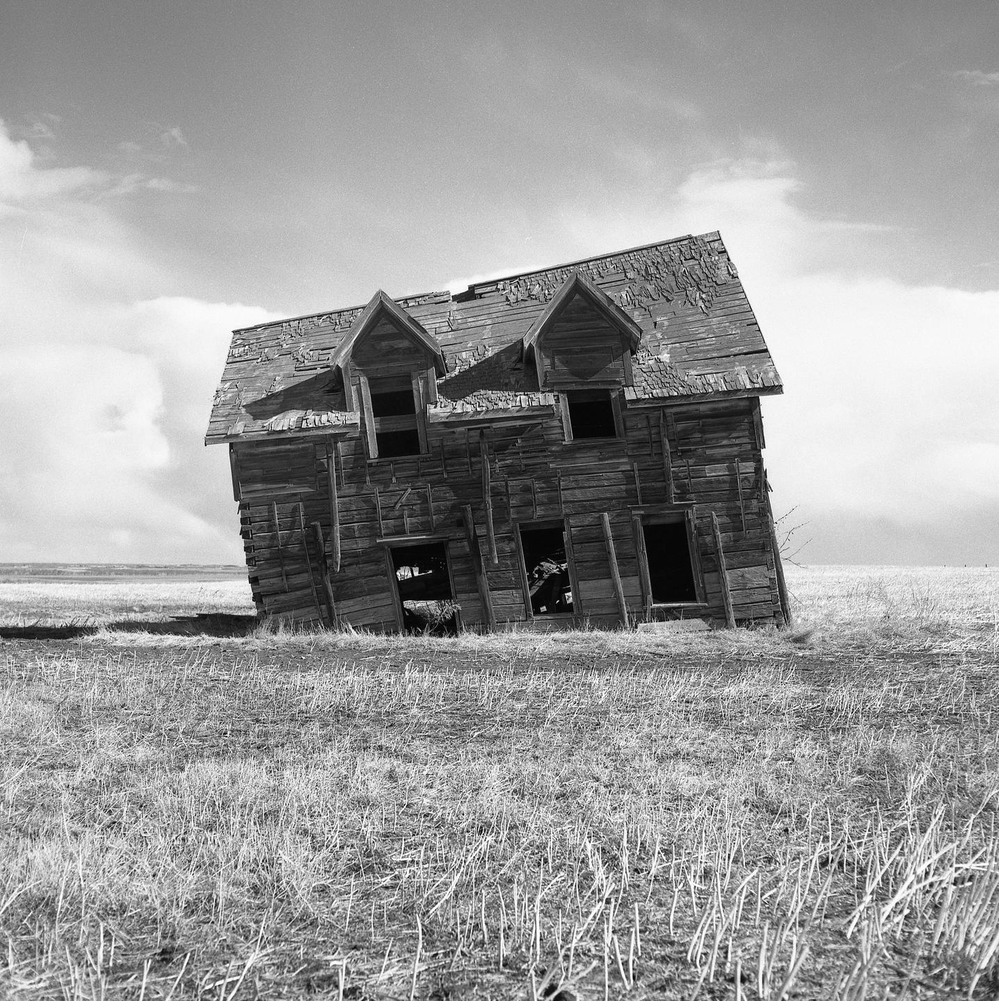 An abandoned wooden house with a leaning facade stands in a field under a cloudy sky.
