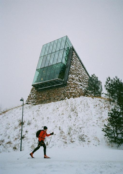 A man cross-country skis in front of a modern building with a glass and stone facade on a snowy hill.