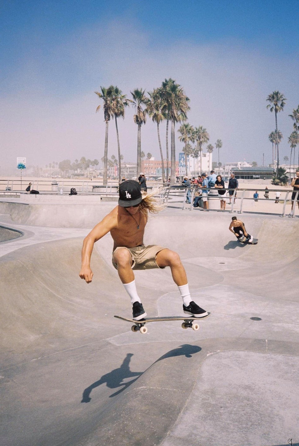 A young man on a skateboard is captured mid-air in a skate park, with palm trees and buildings in the background.