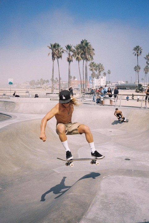 A young man on a skateboard is captured mid-air in a skate park, with palm trees and buildings in the background.
