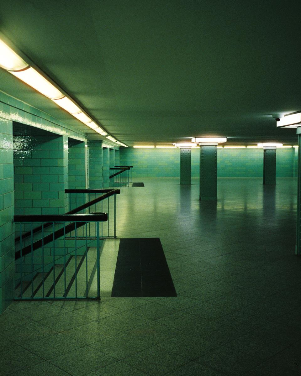A long, empty, green-tiled subway station is illuminated by fluorescent lights.