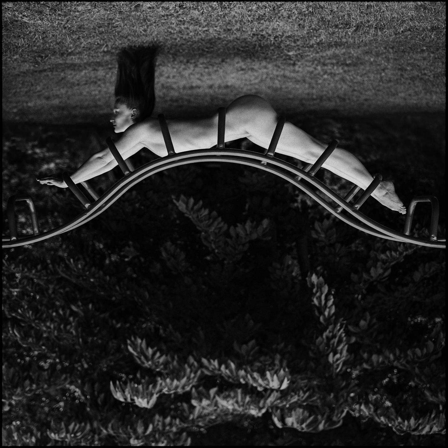 A nude woman is posed on a metal structure, resembling a caterpillar, with her head and hair hanging downwards.