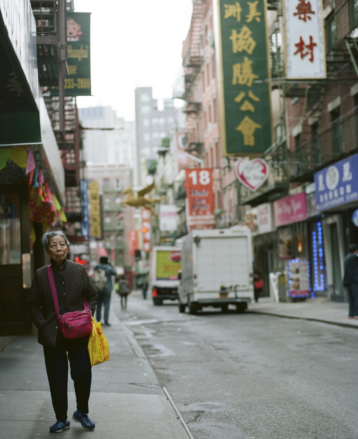 A woman walks on a wet sidewalk in Chinatown, New York, with buildings and trucks in the background.