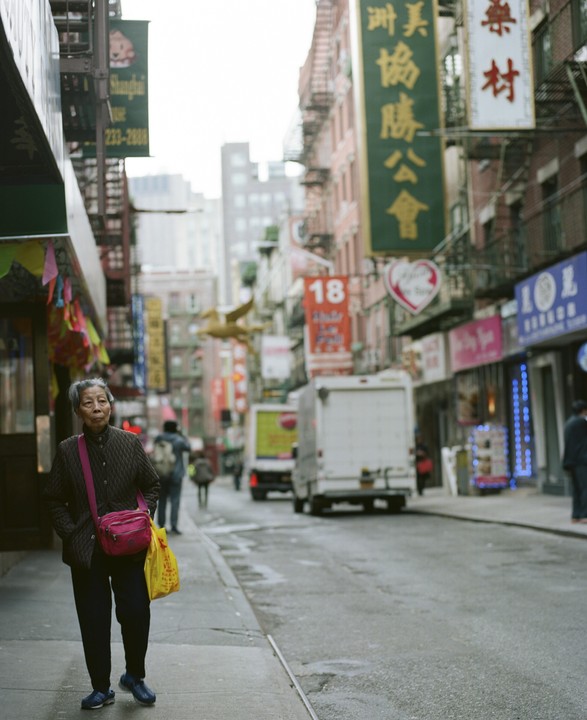 A woman walks on a wet sidewalk in Chinatown, New York, with buildings and trucks in the background.