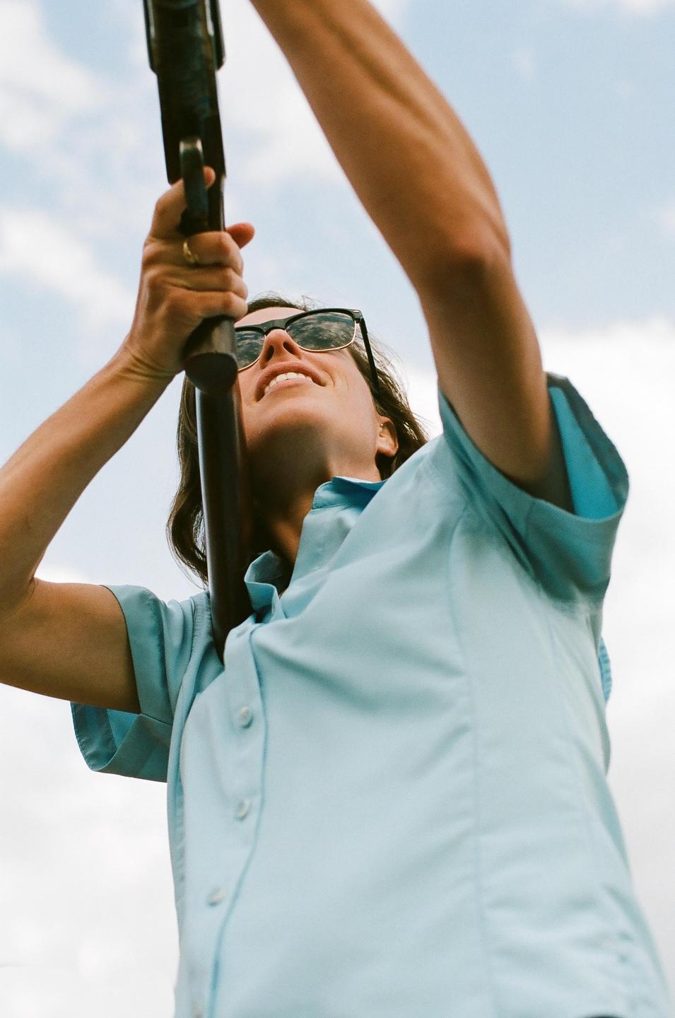 A woman wearing sunglasses and a light blue shirt holds a rifle pointed upwards against a cloudy sky.