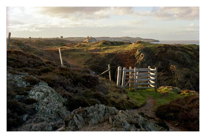 A wooden stile and path lead through a hilly, rocky landscape towards a distant town and the sea under a cloudy sky.