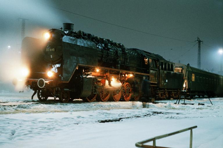 A steam locomotive with its lights on is parked on a snowy track at night.