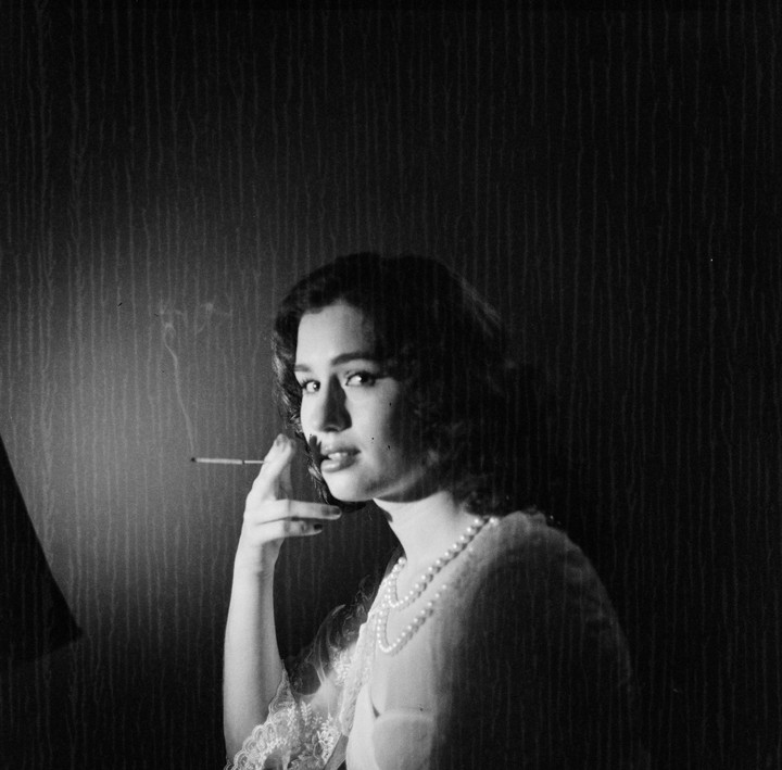 A woman with dark hair and a pearl necklace holds a lit cigarette, looking directly at the viewer.