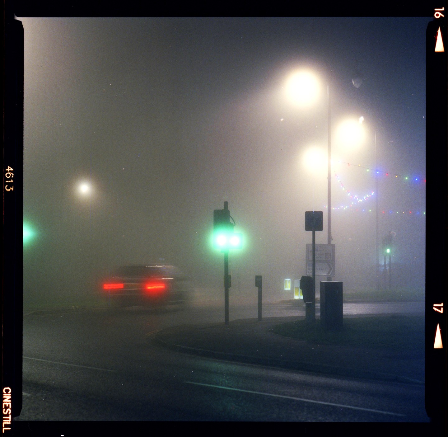 A car with blurred taillights drives through a foggy street at night, illuminated by streetlights and a green traffic signal.