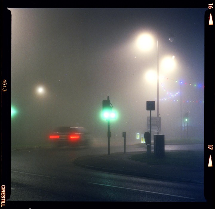 A car with blurred taillights drives through a foggy street at night, illuminated by streetlights and a green traffic signal.
