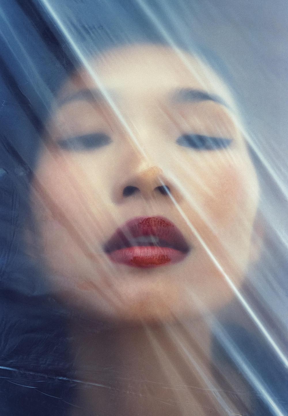 A woman's face with closed eyes and red lips is visible through a translucent, wrinkled material.