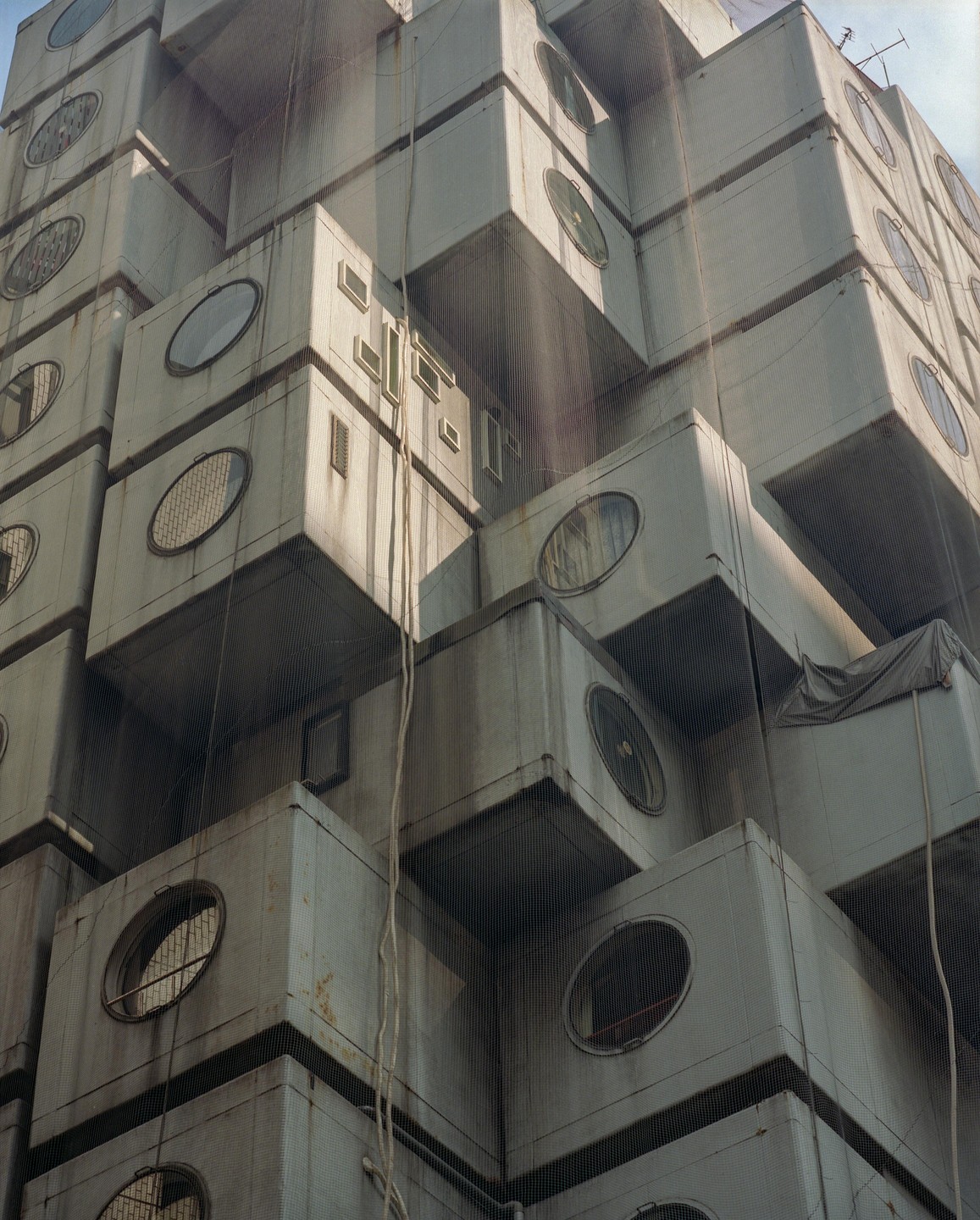 A tall building made of many stacked rectangular modules with round windows is seen from below.
