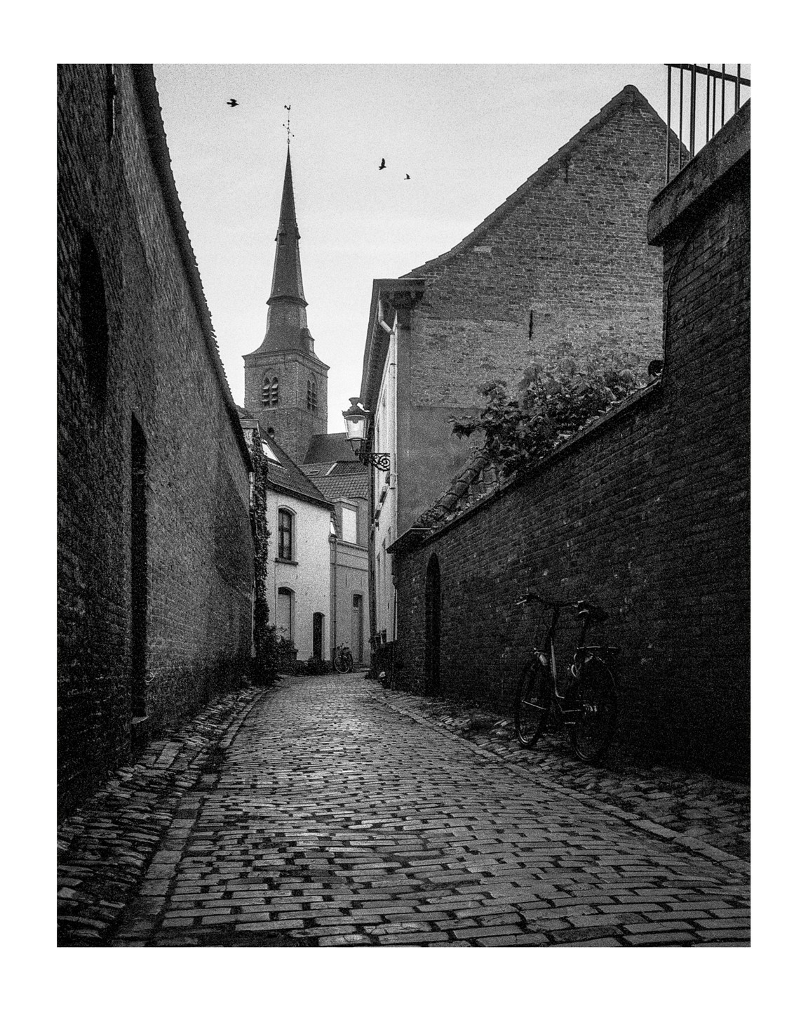 A narrow cobblestone street with brick buildings on either side leads to a church steeple in the distance.