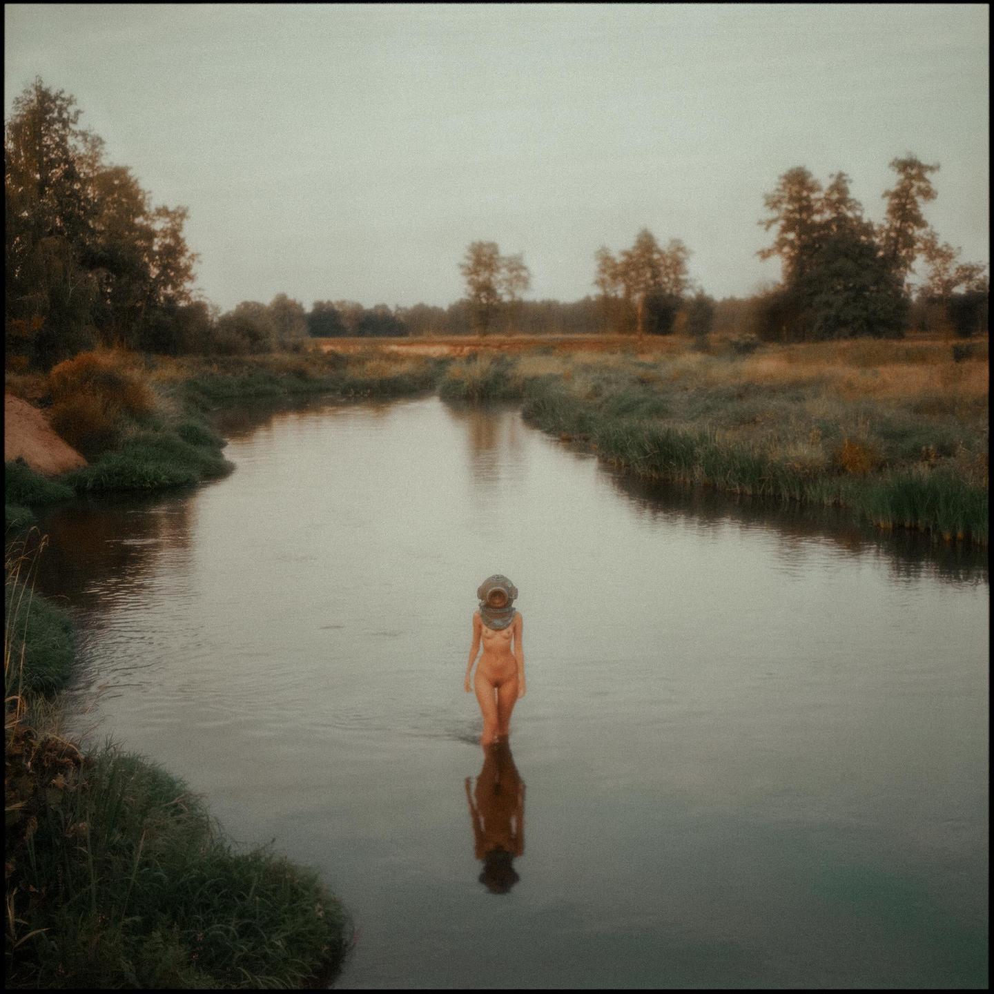 A nude woman wearing a diving helmet stands in a river surrounded by overgrown banks and trees.