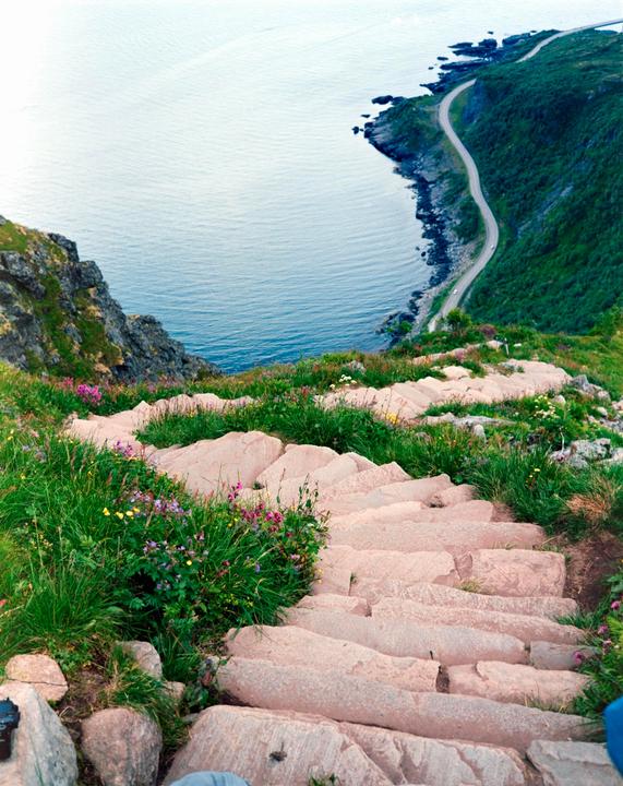 A winding stone staircase descends a grassy hillside towards a coastal road and the ocean.