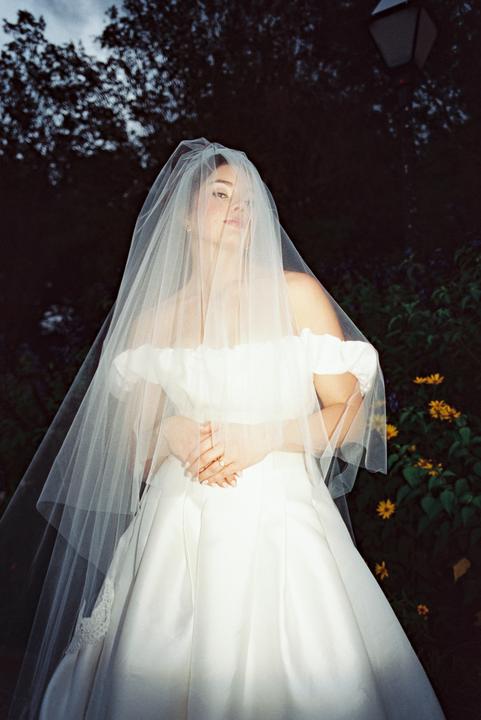 A woman in a white wedding dress and veil stands in a garden at dusk.