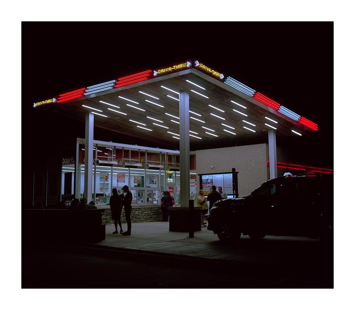 A drive-thru restaurant with neon lights is illuminated at night, with people and a car visible.