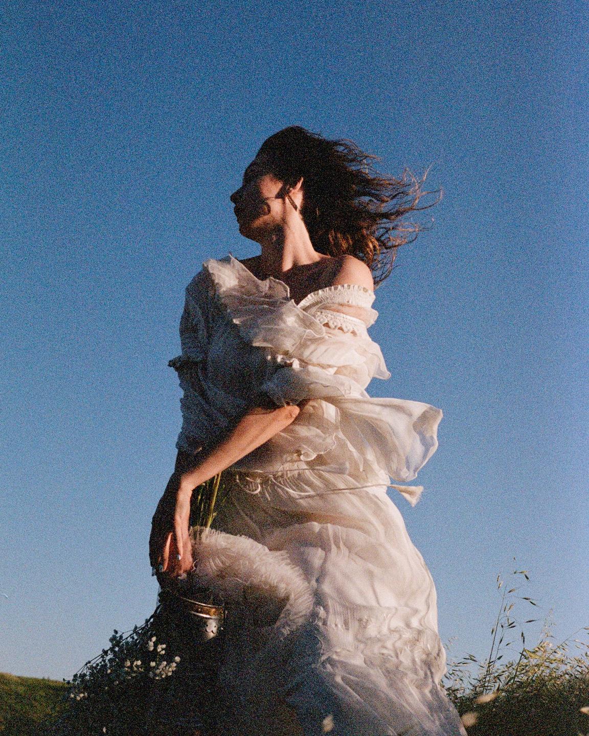 A woman in a white dress stands in a field with her hair blowing in the wind under a clear blue sky.