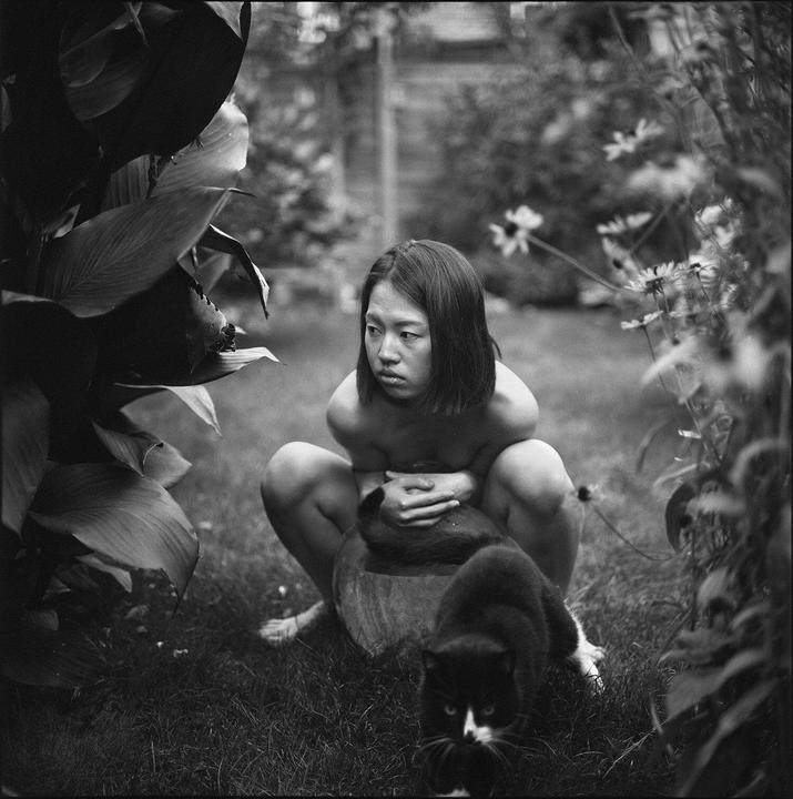 A woman crouches in a garden with a black and white cat in the foreground.