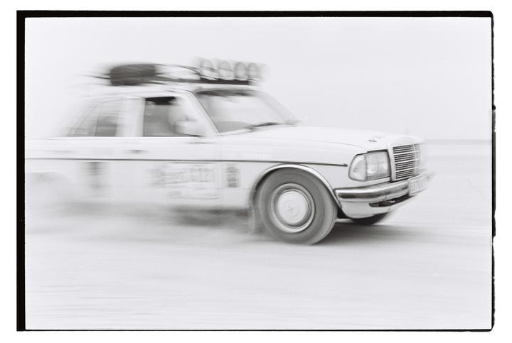 A white car with a roof rack is captured in motion, blurring the background.