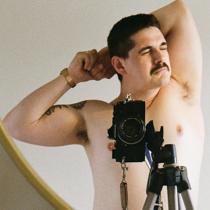 A man with a mustache stretches his arms behind his head while taking a self-portrait in a mirror.