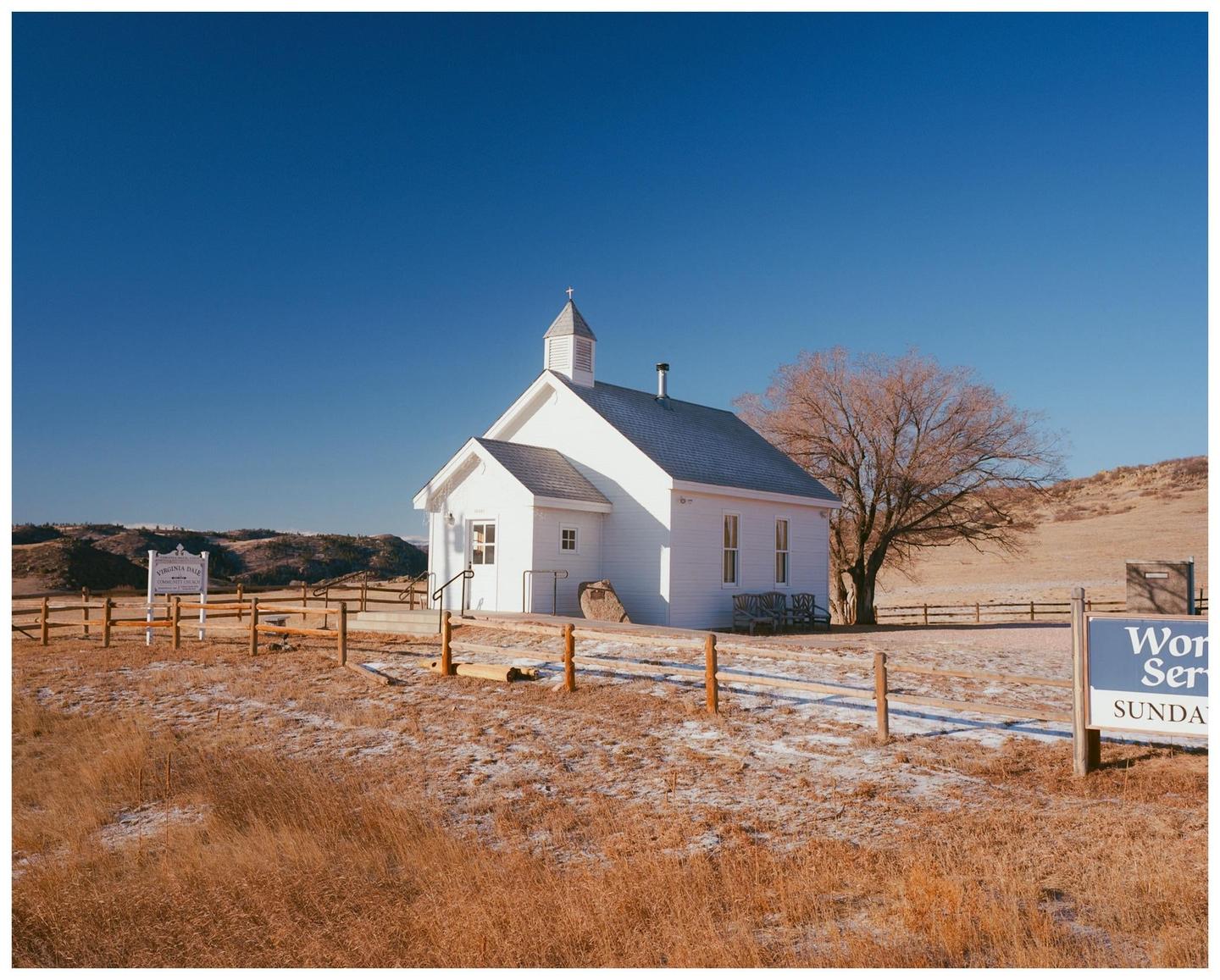 A white church with a steeple stands in a field with a bare tree under a clear blue sky.