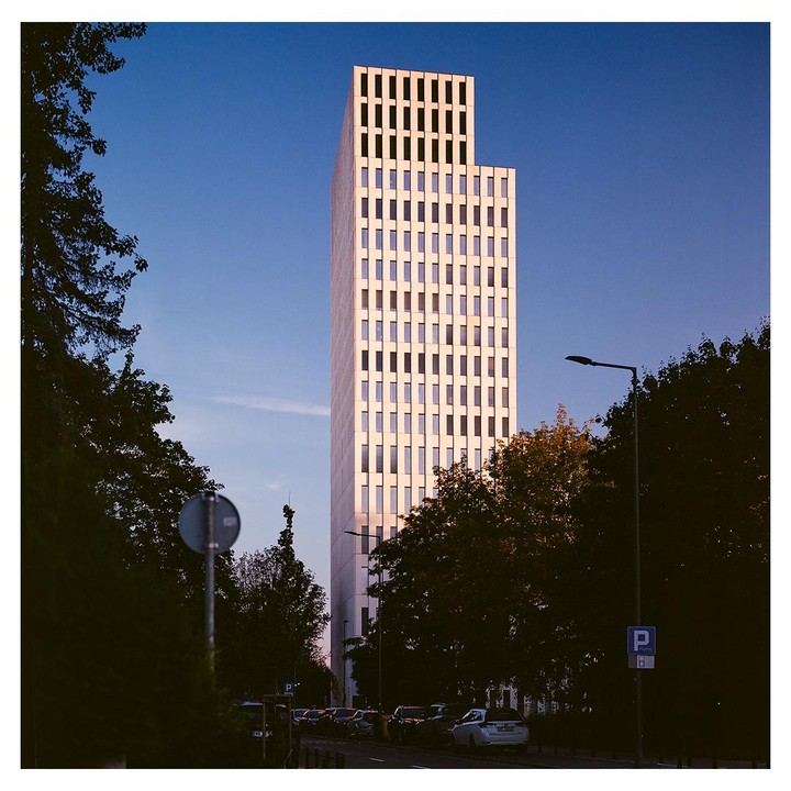 A tall, modern building with many windows stands against a clear blue sky, with trees and parked cars in the foreground.