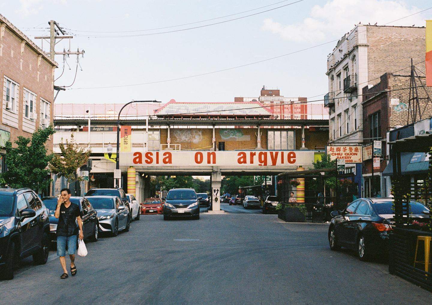 A man walks on a street with parked cars and buildings under a sign that reads "asia on argyle".