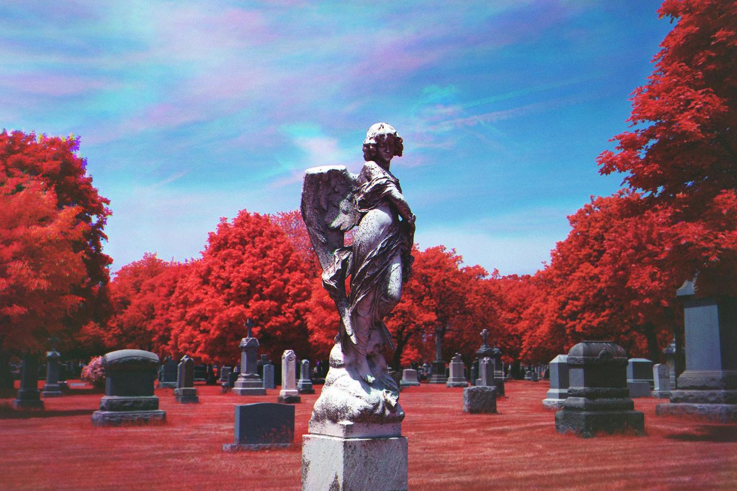 An angel statue stands in a cemetery with red trees and red grass under a blue sky.
